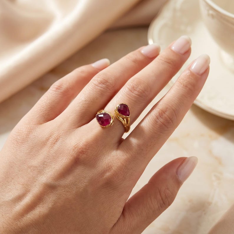 May include: An open-style gold ring featuring two teardrop-shaped, faceted deep red gemstones. The band has a textured, organic finish and is positioned on the finger, showcasing the two stones separated by a gap in the center.