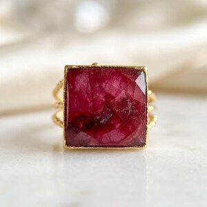 May include: A gold ring featuring a large, square-cut, deep red gemstone with a faceted surface. The band of the ring has a textured, twisted metal design, providing a contrast to the smooth, flat edges of the central square stone setting.