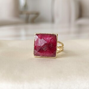 May include: A gold ring featuring a large, square-cut, faceted deep magenta gemstone set in a thin, textured gold frame. The band of the ring consists of three delicate, twisted gold wires that converge into a single shank at the back.