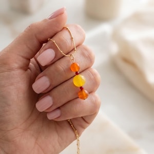 May include: A gold chain necklace featuring a vertical pendant arrangement of three round beads in orange, yellow, and orange, separated by two tiny red spacer beads. The delicate gold chain has small evenly spaced gold beads along its length.