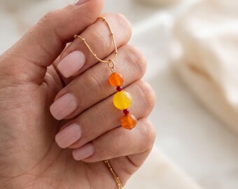 May include: A gold chain necklace featuring a vertical pendant arrangement of three round beads in orange, yellow, and orange, separated by two tiny red spacer beads. The delicate gold chain has small evenly spaced gold beads along its length.
