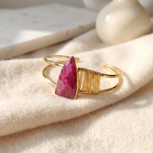 May include: A gold-toned cuff bracelet featuring a triangular, faceted deep pink gemstone as the central focal point. The band is constructed from two parallel wires, with one side accented by a decorative section of tightly coiled gold wire.