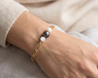 May include: A gold-toned chain bracelet featuring a central arrangement of three spherical beads. The design includes two smaller white beads flanking a single larger dark grey bead, all integrated into a series of elongated oval metal links.