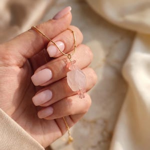 Könnte beinhalten: Eine goldene Halskette mit einem zentralen Anhänger, bestehend aus einer großen, facettierten, blassrosa Quarzperle, die von zwei kleineren, durchscheinenden rosa Kristallperlen flankiert wird und eine feine Anordnung an einer dünnen Kette bildet.