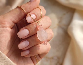 May include: A gold chain necklace featuring a central pendant composed of a large, faceted, pale pink quartz bead flanked by two smaller, translucent pink crystal beads, creating a delicate and balanced arrangement on a thin metallic chain.