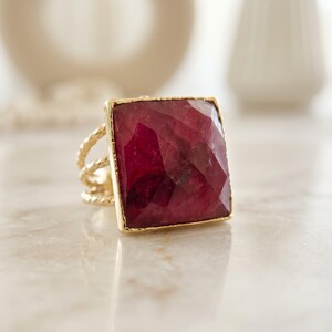 May include: A gold ring featuring a large, square-cut, deep red gemstone with a faceted surface. The band of the ring is constructed from three thin, twisted gold wires that converge into a single shank at the base of the setting.