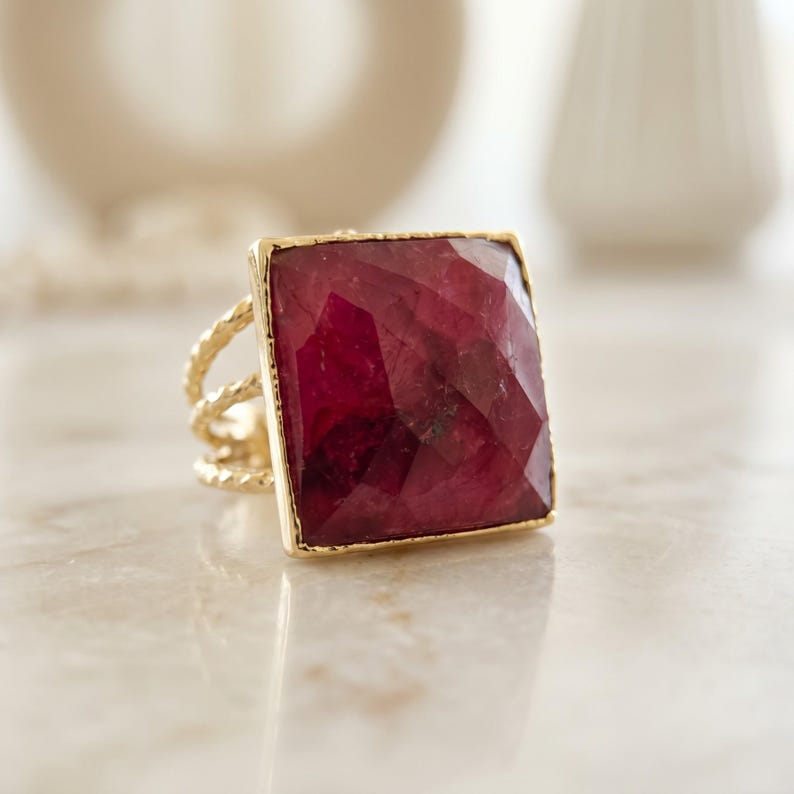 May include: A gold ring featuring a large, square-cut, deep red gemstone with a faceted surface. The band of the ring is constructed from three thin, twisted gold wires that converge into a single shank at the base of the setting.