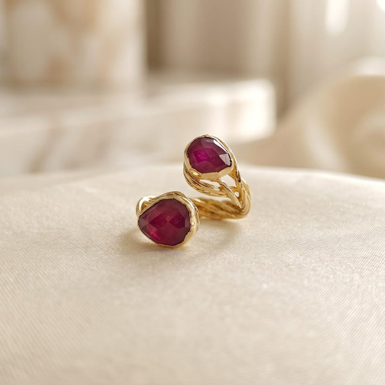 May include: A gold open-style ring featuring two faceted, deep red, pear-shaped stones. The band has a twisted, textured metal design that connects the two stones at opposite ends of the open gap in the center of the ring.