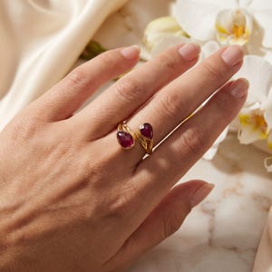 May include: A gold open-style ring featuring two teardrop-shaped, deep red faceted gemstones. The band has a textured, organic finish that tapers toward the stones, which are positioned at the open ends of the ring.