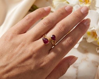 May include: A gold open-style ring featuring two teardrop-shaped, deep red faceted gemstones. The band has a textured, organic finish that tapers toward the stones, which are positioned at the open ends of the ring.