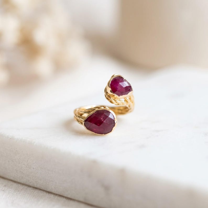 May include: A gold open-style ring featuring two pear-shaped, faceted deep red rubies set at each end. The band has a textured, twisted rope-like design that connects the two stones, creating a bypass silhouette with a polished finish.