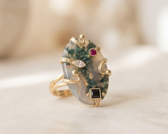 May include: A gold ring featuring a large oval moss agate stone with green inclusions. The setting includes a small round red gemstone, a marquise-cut white stone, and a square black stone, all held by delicate gold prongs on a split band.