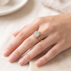 May include: A gold-toned ring featuring a square-shaped, textured white druzy stone centrepiece. The band is thin and open-ended, with a small gold sphere at the end of the wire, creating a bypass style design that sits on the finger.