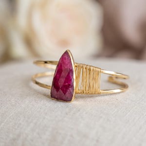 May include: A gold cuff bracelet featuring a faceted, triangular-shaped deep pink gemstone. The metal band is accented with delicate gold wire wrapping that secures the stone to the open-ended frame, creating a structured and layered appearance.