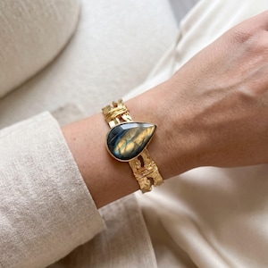 Può includere: Un bracciale rigido color oro con una pietra preziosa di labradorite a forma di goccia, caratterizzata da riflessi blu e dorati. La fascia in metallo presenta una finitura martellata e testurizzata, arricchita da sottili fili avvolti.