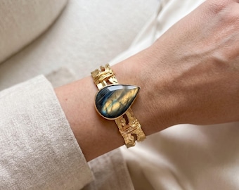 May include: A gold-toned cuff bracelet featuring a central teardrop-shaped labradorite gemstone with blue and gold iridescence. The metal band has a textured, hammered finish and is accented with thin wire wrapping details around the stone.