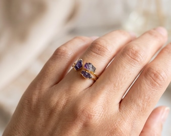 May include: A gold-toned open ring featuring three small, raw, purple amethyst stones set along the band. The stones have an irregular, natural shape and are positioned at the open ends of the thin, metallic ring band, creating a layered look.