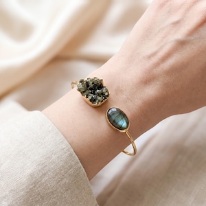 May include: A gold-toned open cuff bracelet featuring two distinct stones. One end displays a dark, textured, crystalline cluster, while the other end holds a smooth, oval-shaped labradorite stone with a visible blue-green iridescent sheen.