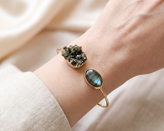May include: A gold-toned open cuff bracelet featuring two distinct stones. One end displays a dark, textured, crystalline cluster, while the other end holds a smooth, oval-shaped labradorite stone with a visible blue-green iridescent sheen.