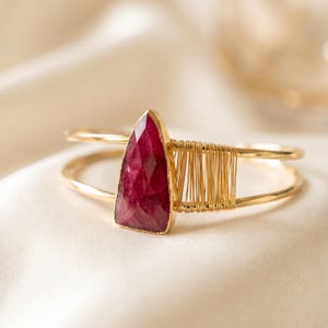 May include: A gold-tone cuff bracelet featuring a single, triangular, faceted deep pink gemstone. The stone is set on a thin metal band, with one side of the band wrapped in fine gold-tone wire, creating a textured, layered appearance.
