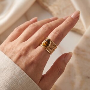 May include: A gold-toned ring featuring a teardrop-shaped tiger's eye gemstone set in the centre. The band of the ring is constructed from multiple thin, layered gold-toned wires that wrap around the finger, creating a textured, open-work design.