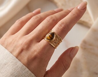 May include: A gold-toned ring featuring a teardrop-shaped tiger's eye gemstone set in the centre. The band of the ring is constructed from multiple thin, layered gold-toned wires that wrap around the finger, creating a textured, open-work design.
