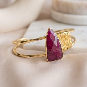 May include: A gold-toned cuff bracelet featuring a faceted, teardrop-shaped deep pink gemstone as the central focal point. The metal band is open-ended and includes a section of decorative gold wire wrapping adjacent to the stone.
