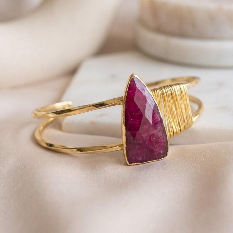 May include: A gold-toned cuff bracelet featuring a faceted, teardrop-shaped deep pink gemstone as the central focal point. The metal band is open-ended and includes a section of decorative gold wire wrapping adjacent to the stone.
