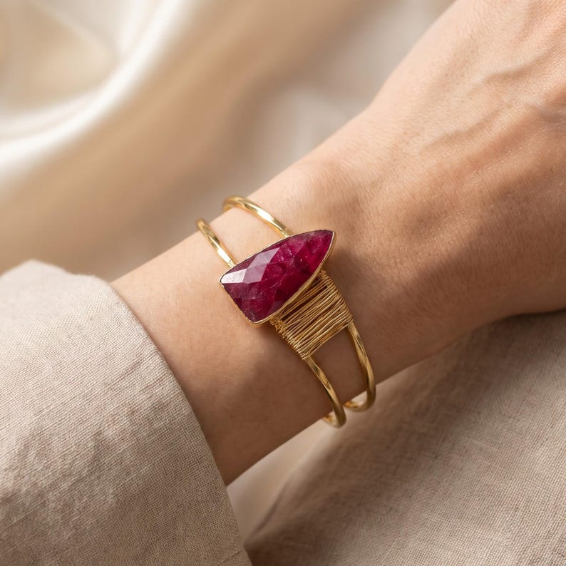 May include: A gold-toned cuff bracelet featuring a double-band design and a faceted, triangular-shaped deep pink gemstone. The stone is secured by thin gold wire wrapping that extends along one side of the metal band structure.