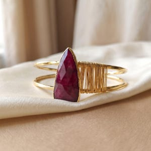 May include: A gold-toned cuff bracelet featuring a faceted, teardrop-shaped deep red gemstone as the centrepiece. The band is constructed from two thin metal wires, with one side wrapped in fine gold-toned wire for a textured detail.