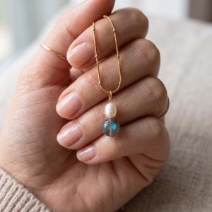 May include: A gold chain necklace featuring a delicate beaded texture, adorned with a single white oval pearl pendant positioned directly above a round, grey labradorite gemstone that displays a subtle iridescent blue flash of light.