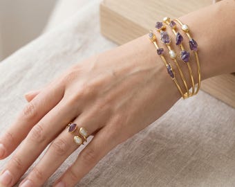 May include: A set of four gold-toned bangles and an open-style ring, each featuring raw purple amethyst stones and small white pearls. The jewelry pieces are crafted with thin, polished metal bands that highlight the natural texture of the gems.