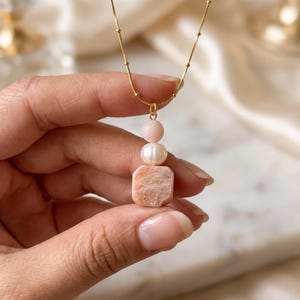 May include: A gold-toned chain necklace featuring a pendant composed of a small round pale pink bead, a white oval pearl, and a square-shaped peach-coloured stone with a shimmering, marbled texture, all suspended vertically from the delicate chain.