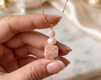 May include: A gold-toned chain necklace featuring a pendant composed of a small round pale pink bead, a white oval pearl, and a square-shaped peach-coloured stone with a shimmering, marbled texture, all suspended vertically from the delicate chain.