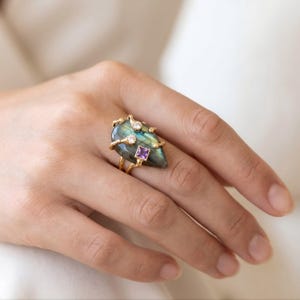 May include: A gold ring featuring a large, pear-shaped labradorite gemstone with a distinct blue and green iridescent sheen. The setting includes two small, round, clear stones and one square-cut purple stone, all mounted on a textured gold band.