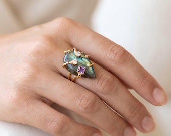May include: A gold ring featuring a large, pear-shaped labradorite gemstone with a distinct blue and green iridescent sheen. The setting includes two small, round, clear stones and one square-cut purple stone, all mounted on a textured gold band.