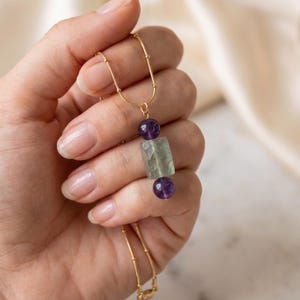 May include: A gold-toned chain necklace featuring a central pendant composed of a rectangular, faceted pale green stone flanked by two small, round, deep purple amethyst beads. The delicate metal chain connects to the pendant via a small jump ring.