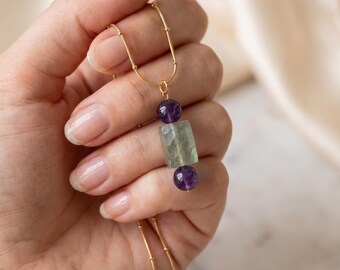 May include: A gold-toned chain necklace featuring a central pendant composed of a rectangular, faceted pale green stone flanked by two small, round, deep purple amethyst beads. The delicate metal chain connects to the pendant via a small jump ring.