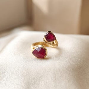May include: A gold open ring featuring two pear-shaped, faceted deep red rubies set at each end. The band has a textured, organic surface that resembles twisted metal, connecting the two gemstone settings in a bypass style design.