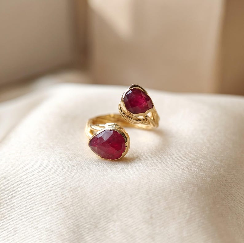 May include: A gold open ring featuring two pear-shaped, faceted deep red rubies set at each end. The band has a textured, organic surface that resembles twisted metal, connecting the two gemstone settings in a bypass style design.