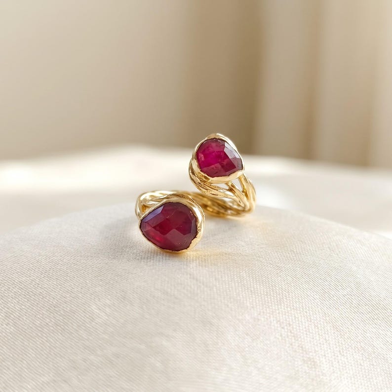 May include: A gold bypass ring featuring two faceted, pear-shaped deep red rubies set at each end. The band is crafted from textured, twisted gold wire that wraps around the finger, showcasing a polished finish and vibrant gemstone colors.