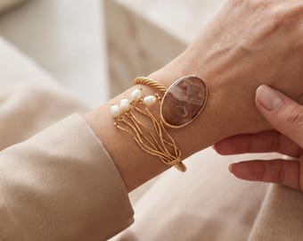 May include: A gold-toned cuff bracelet featuring a twisted metal band, a large oval brown and beige patterned stone, and a decorative cluster of four small white pearls attached to thin, wavy gold-toned metal wires extending from the main band.