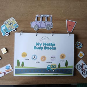 Könnte beinhalten: Ein My Maths Busy Books Lernordner mit Metallringen, der eine Straßengrafik und pädagogische Symbole zeigt. Um den Ordner herum befinden sich verschiedene ausgeschnittene Formen, darunter ein blauer Lastwagen, ein lila Auto, ein Saftbecher und eine Ente.