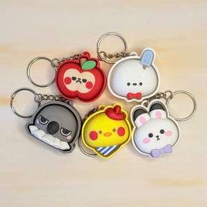 May include: Five colorful silicone keychains featuring cartoon characters: a red apple with a face, a white round creature with a blue popsicle, a gray koala, a yellow chick with a red hat, and a white rabbit with a purple bow tie.