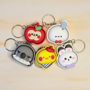 May include: A collection of five colorful silicone keychains featuring various characters: a red apple, a white round creature with a blue popsicle, a gray koala, a yellow chick with a red hat, and a white rabbit with a purple bow tie.