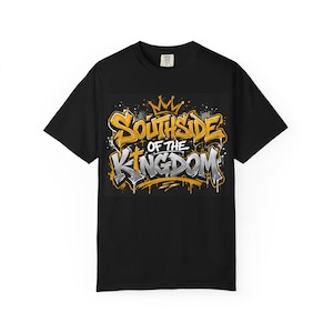 以下が含まれることがあります： 「SOUTHSIDE OF THE KINGDOM」というグラフィティスタイルの文字が中央にプリントされた黒の半袖Tシャツ。文字の上には金色の王冠が描かれており、金と銀の文字にはペンキが垂れたようなアクセントが施されています。