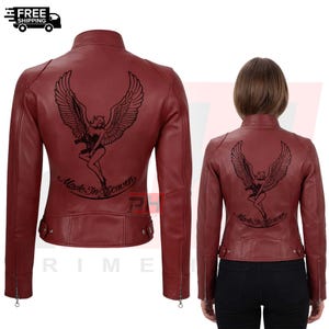 May include: A dark red jacket featuring a black graphic on the back of a winged figure holding a firearm, with the text Made In Heaven printed below. The jacket has a stand-up collar, long sleeves with zippered cuffs, and side waist tabs.