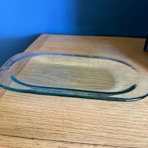 May include: A clear, oval-shaped glass tray with a slightly raised rim and a smooth, transparent surface. The glass has a subtle greenish tint along the edges, reflecting light across its flat, elongated base and rounded, curved perimeter.