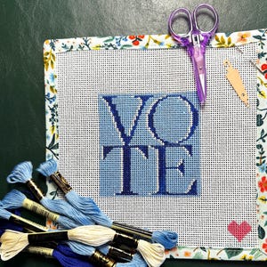 May include: A needlepoint canvas with the word "VOTE" stitched in blue, surrounded by a floral patterned border. Includes blue embroidery floss, purple scissors, and a small tag. A pink heart is stitched in the lower right corner.