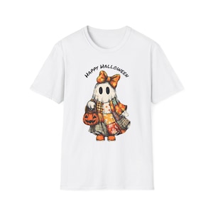 Puede incluir: Una camiseta blanca de manga corta con el gráfico de un fantasma que lleva un lazo naranja grande y un vestido de retazos. El fantasma sostiene un cubo de calabaza naranja. El texto "HAPPY HALLOWEEN" está impreso en un arco negro sobre la figura.